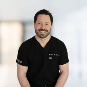 Sean Langton, DMD, is smiling while wearing black medical scrubs with the text "Meet Our Doctors." He is standing against a blurred, bright background at Belmont Dental.