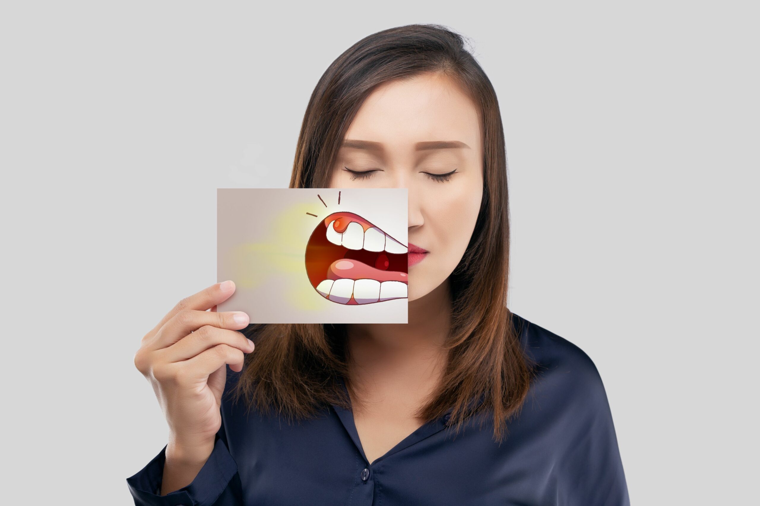 A woman with closed eyes holds a card in front of her mouth. The card depicts an open mouth with a painful tooth highlighting dental discomfort.