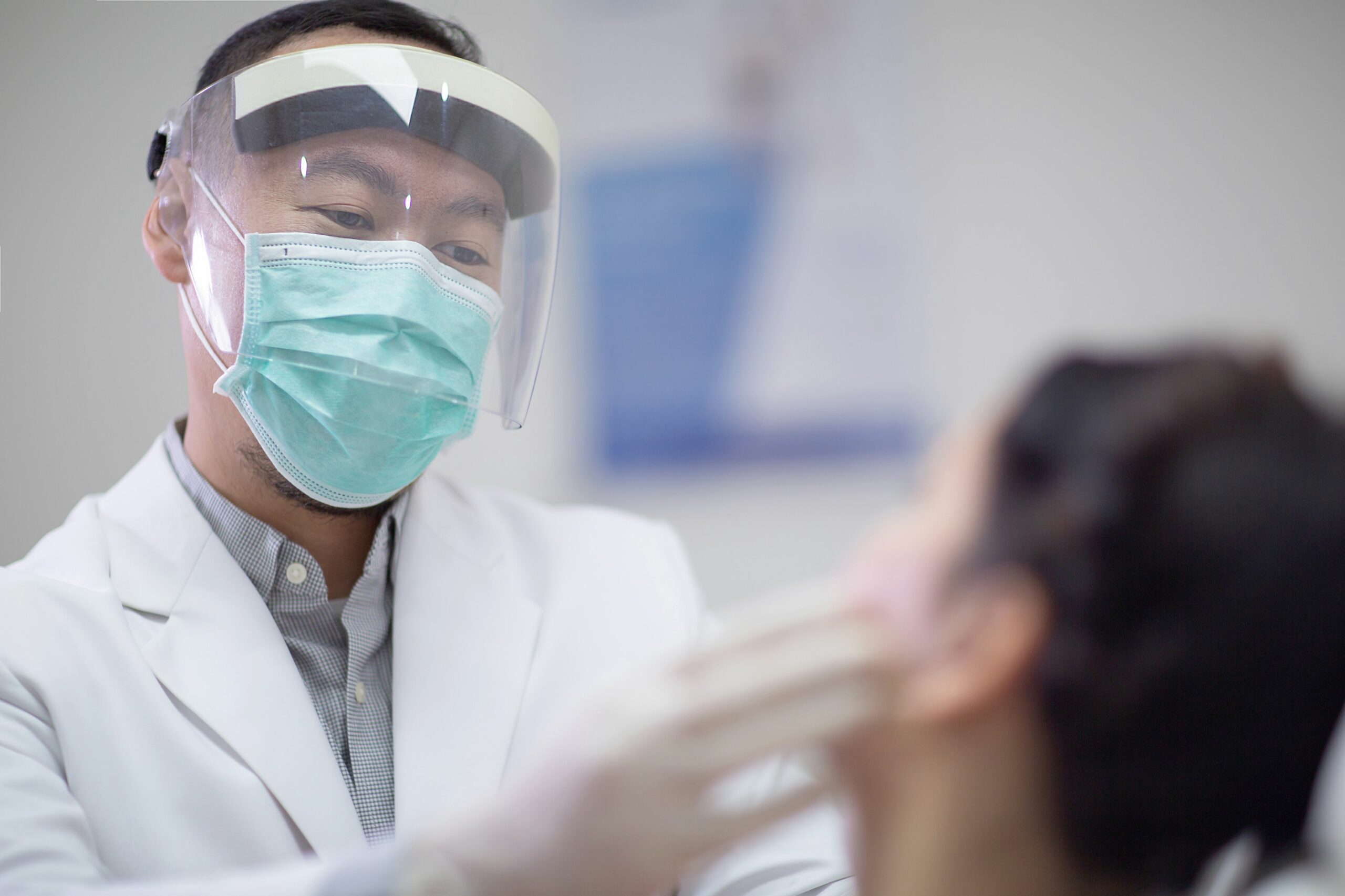 A medical professional wearing a face shield and mask examines a patient's face in a clinical setting.