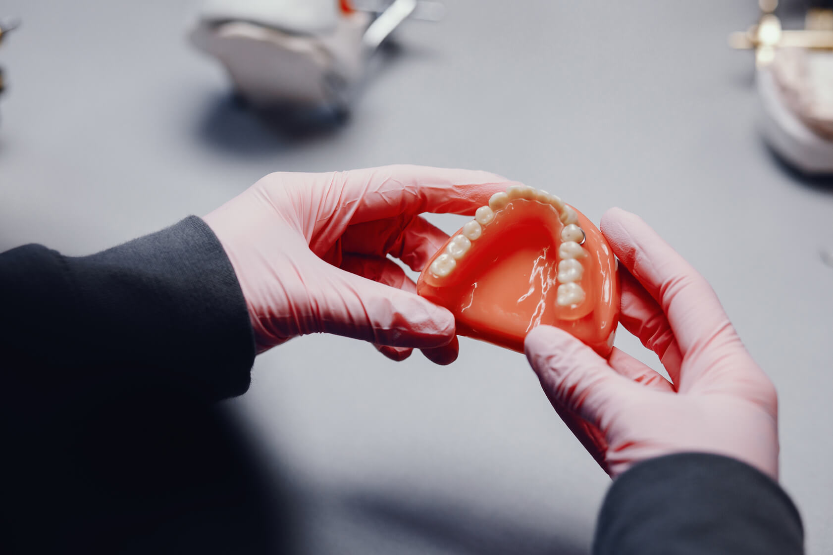 A person wearing pink gloves holds a dental prosthesis, displaying artificial teeth, with other dental models in the background.