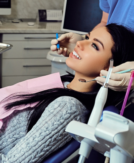 A woman smiles while sitting in a dental chair as a dentist, wearing gloves, examines her teeth using dental tools.
