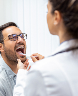 A male patient with glasses opens his mouth as a healthcare worker, seen from behind, uses a swab to take a sample from his throat.