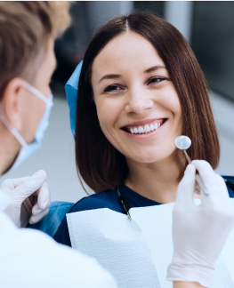 A woman sits in a dental chair, smiling at a dentist wearing a mask and gloves, who is holding a dental mirror.