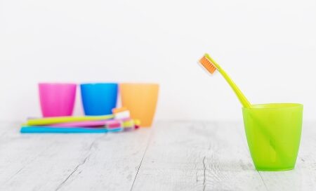 A yellow toothbrush with an orange head stands in a green plastic cup. In the background, several colorful cups and toothbrushes lie on a white surface.