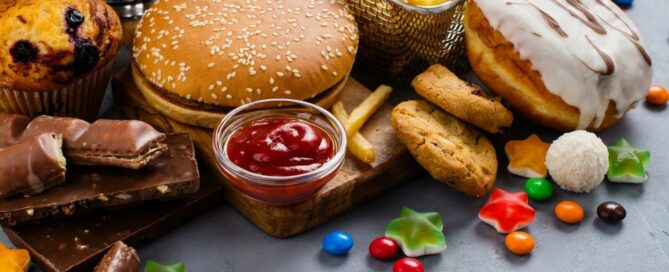 A variety of fast foods and sweets, including a hamburger, fries, nuggets, donuts, cookies, chocolate bars, candies, a muffin, and a soda, displayed on a gray surface.