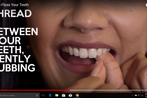 A person demonstrates how to floss their teeth, with text on the image saying "THREAD BETWEEN YOUR TEETH, GENTLY RUBBING.