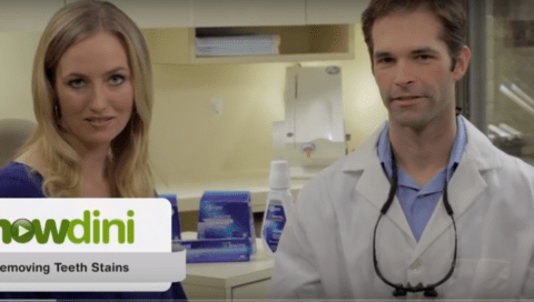 A female and a male are standing together in a dental office. The man is wearing a lab coat and dental loupes. Overlaid text reads, "howdini - Removing Teeth Stains." The video title says, "Teeth Whitening Tips: How to Remove Stains From Teeth.