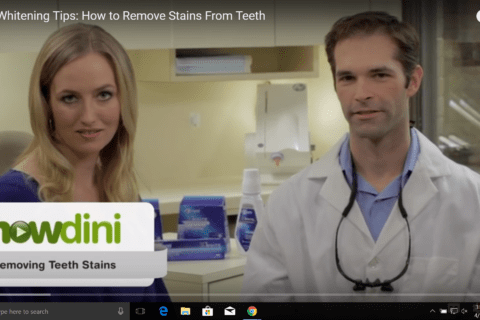 A female and a male are standing together in a dental office. The man is wearing a lab coat and dental loupes. Overlaid text reads, "howdini - Removing Teeth Stains." The video title says, "Teeth Whitening Tips: How to Remove Stains From Teeth.