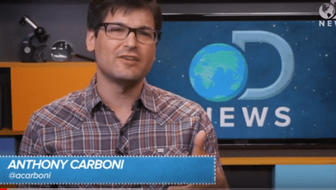 A person wearing glasses and a plaid shirt gives a thumbs-up gesture while speaking in front of a TV screen displaying the text "D NEWS." On-screen text labels them as "Anthony Carboni @acarboni.