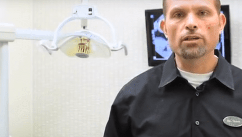Man standing in a dental office, wearing a black shirt with a name badge, a dental light and a monitor behind him.