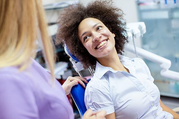 A woman sits smiling in a dental chair while a dental professional stands beside her.