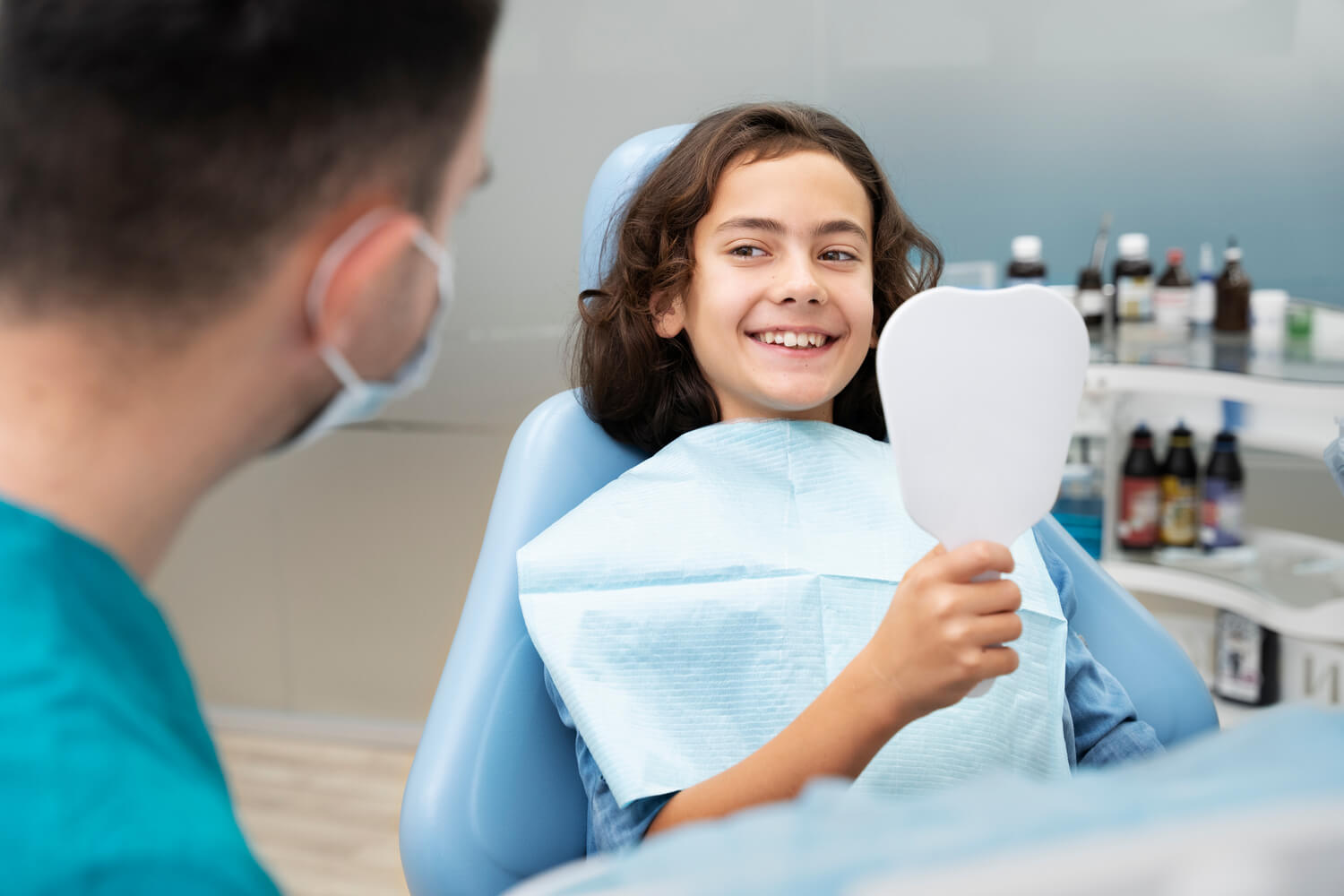 A child in a dental chair smiles while holding a mirror as a dentist in a mask looks on, showcasing their top-notch dental services.