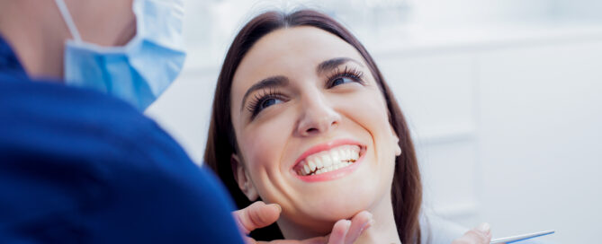 A dentist examines a smiling woman’s teeth while she sits in a dental chair.
