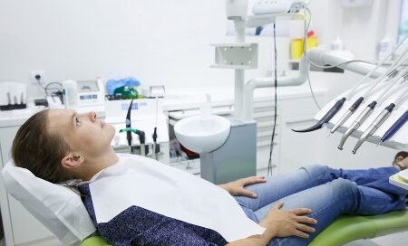 A person lies back in a dental chair with a bib on, looking upwards in a dental office surrounded by various dental instruments.