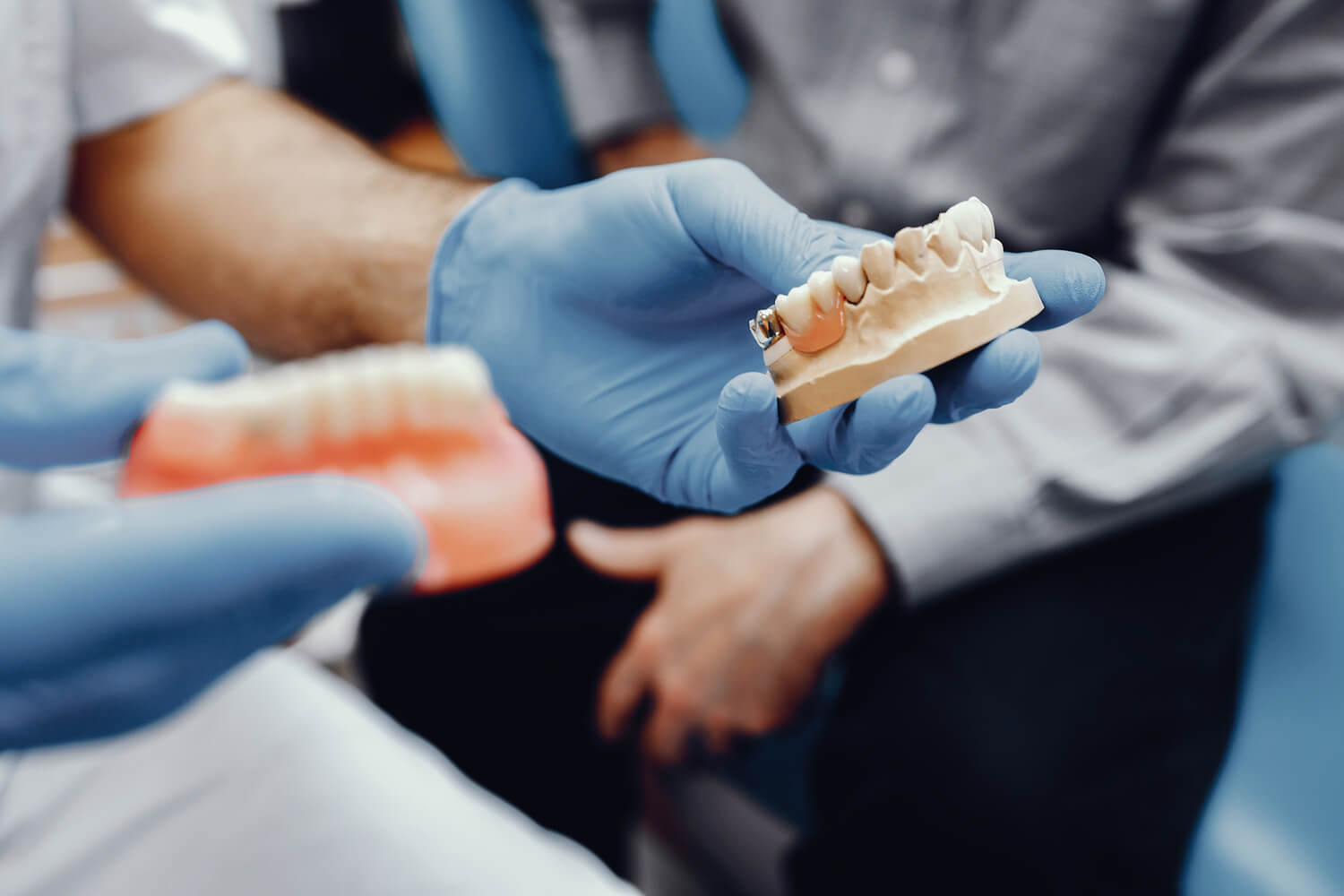 A dentist wearing blue gloves holds two dental models, explaining their details to a seated patient, highlighting the comprehensive dental services available.