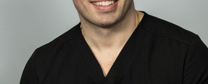 Man in black medical scrubs with embroidered name and title, smiling and seated against a plain background.