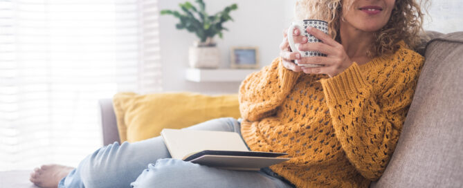 A person with curly hair sits on a couch holding a mug, with an open notebook resting on their lap, possibly preparing notes for an upcoming dental surgery. They are wearing a yellow sweater and jeans. A potted plant is visible in the background.