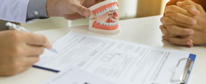 A dentist explains with a dental model while a patient listens attentively, mindful of any allergies. Documents and a clipboard rest on the desk between them.