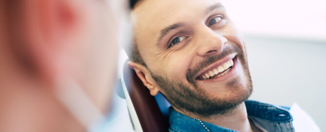Man seated in a dental chair, smiling, wearing a denim shirt, with a dentist blurred in the foreground.