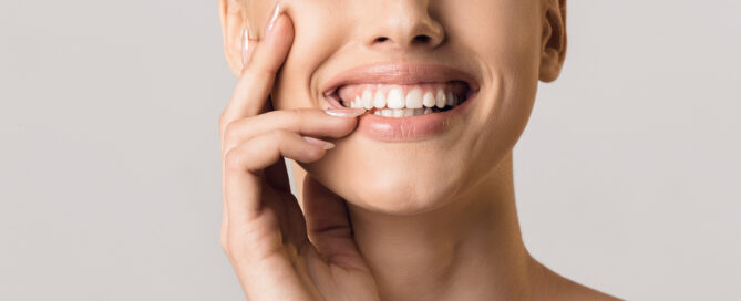 Close-up of a smiling person with a hand touching their face, displaying a set of white teeth. The background is plain and light gray.