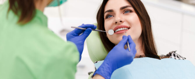 A dentist wearing green scrubs and blue gloves examines a smiling patient's teeth using dental tools while the patient sits in a dental chair.