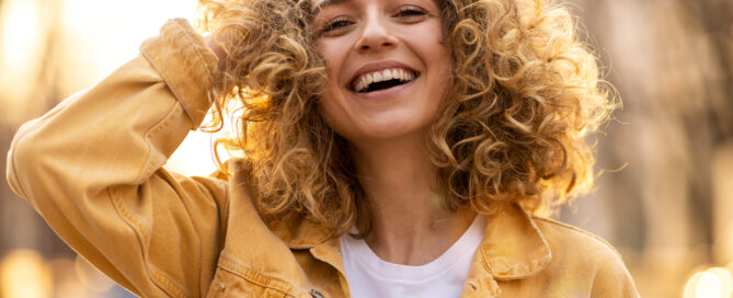 A person with curly hair smiles while touching their hair, revealing the glint of enamel pins on their yellow jacket over a white shirt. The background is blurred, with warm light suggesting it's taken outdoors.