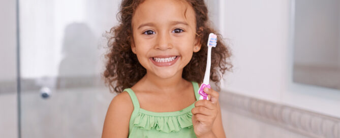 A young girl in a green top holding a toothbrush smiles in a bathroom, embodying essential dental advice for parents.