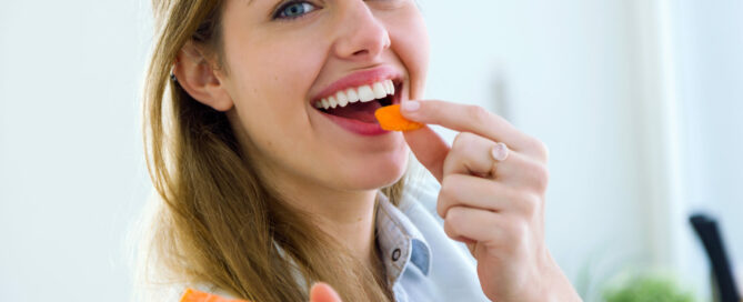 A woman smiles while holding a carrot stick, with another carrot stick raised near her mouth.