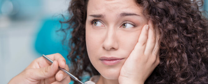 A person with curly hair looks worried, holding their cheek with one hand, while a dentist's hand holding a dental mirror is near their face.