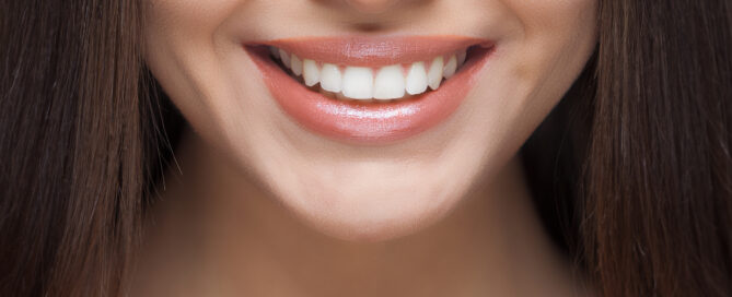 Close-up of a woman's smile with straight, white teeth and glossy lips. The image shows only the lower half of her face and her long, straight brown hair.