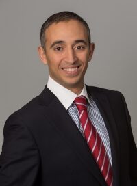 A person with short dark hair, wearing a dark suit, a white shirt, and a red striped tie, is smiling at the camera with a neutral gray background.