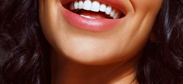 Close-up of a person smiling, showing white teeth and pink lips. The person's hair is dark and wavy, partially visible.