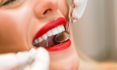 A person with bright red lipstick smiling while a dentist uses a dental mirror and probe to examine their teeth.