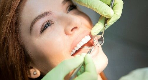 A person is lying back in a dental chair with their mouth open while a dentist in green gloves examines their teeth using dental instruments.