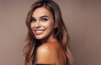 Smiling woman with long, light brown hair wearing an off-the-shoulder top against a neutral background, looking slightly to the side.