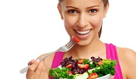 A woman smiles while holding a fork with a cherry tomato next to a bowl of salad consisting of lettuce, tomatoes, cucumbers, and other greens.