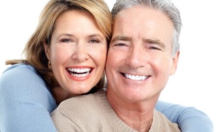 A smiling middle-aged woman with blond hair hugs a smiling middle-aged man with gray hair from behind. They both wear light-colored sweaters and look directly at the camera.