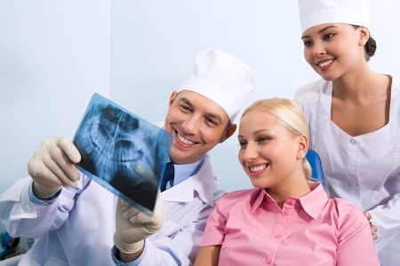 A dentist shows a dental X-ray to a smiling patient. Another dental assistant is also present in the room.