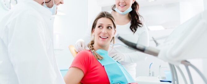 A pregnant woman sits in a dental chair, smiling at a male dentist and a female dental assistant who are engaging with her.