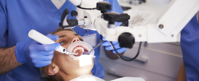 A patient undergoes a dental procedure while lying in a chair. Two dental professionals in blue scrubs and gloves use instruments and an overhead microscope to perform the treatment.