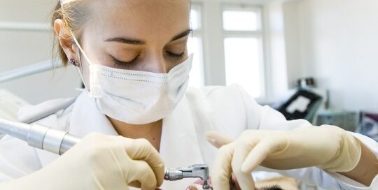 A dental professional wearing a mask, protective eyewear, and gloves, is performing a dental procedure on a patient lying down with their mouth open.