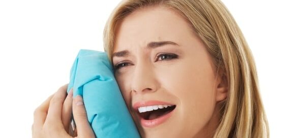 A woman holds an ice pack to her cheek with a pained expression, indicating discomfort or toothache. She has light brown hair and is facing slightly to the left.