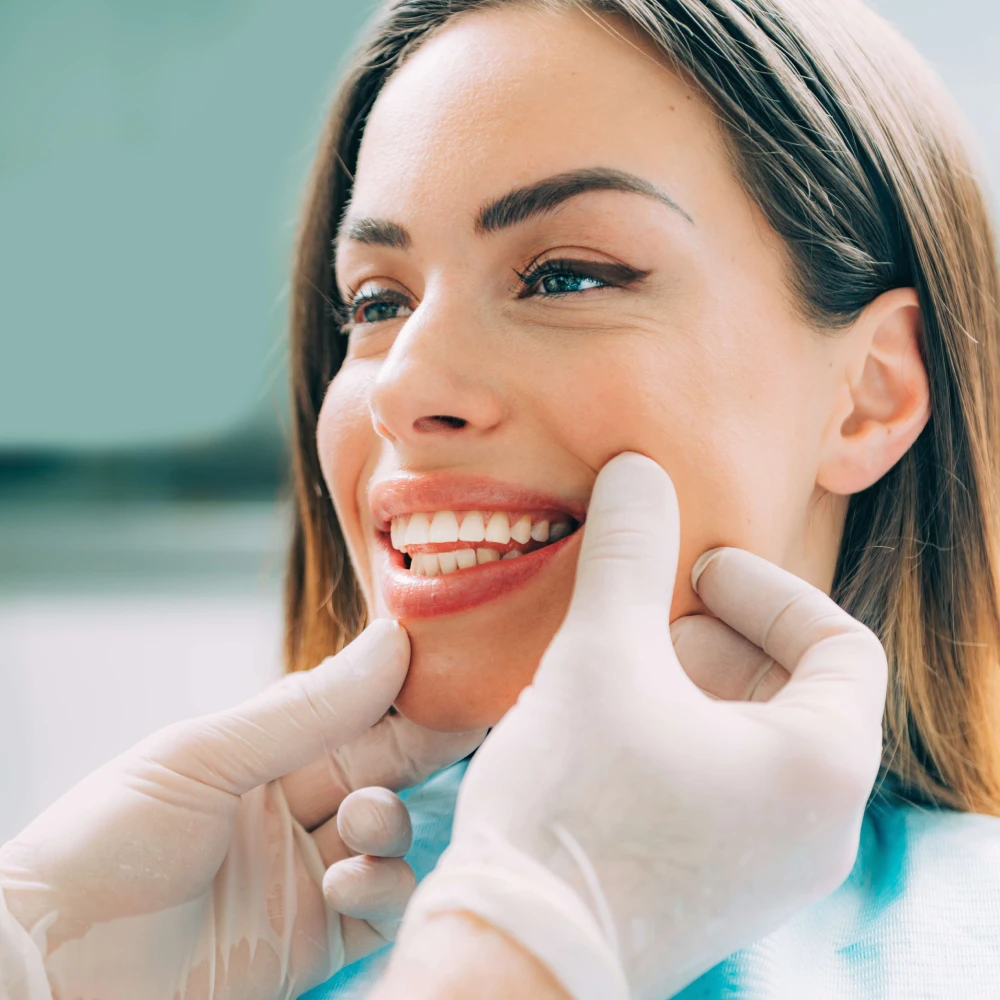 A dentist wearing gloves examines a smiling patient's teeth, gently holding the patient's cheeks, and explaining the benefits of sealants for long-term dental health.
