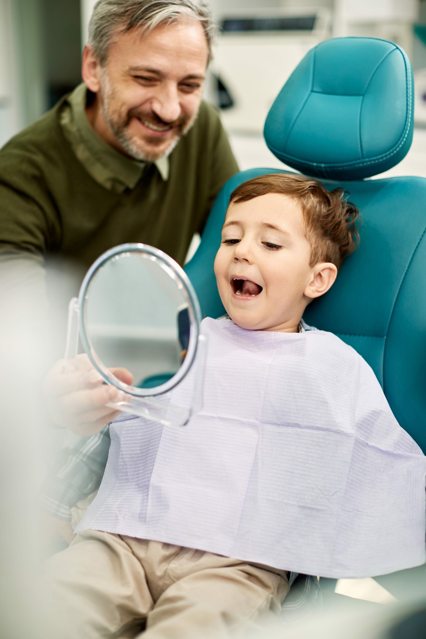 A man shows a young boy his reflection in a handheld mirror while sitting in a dentist chair. The boy looks at his reflection with an open mouth. Both are smiling.
