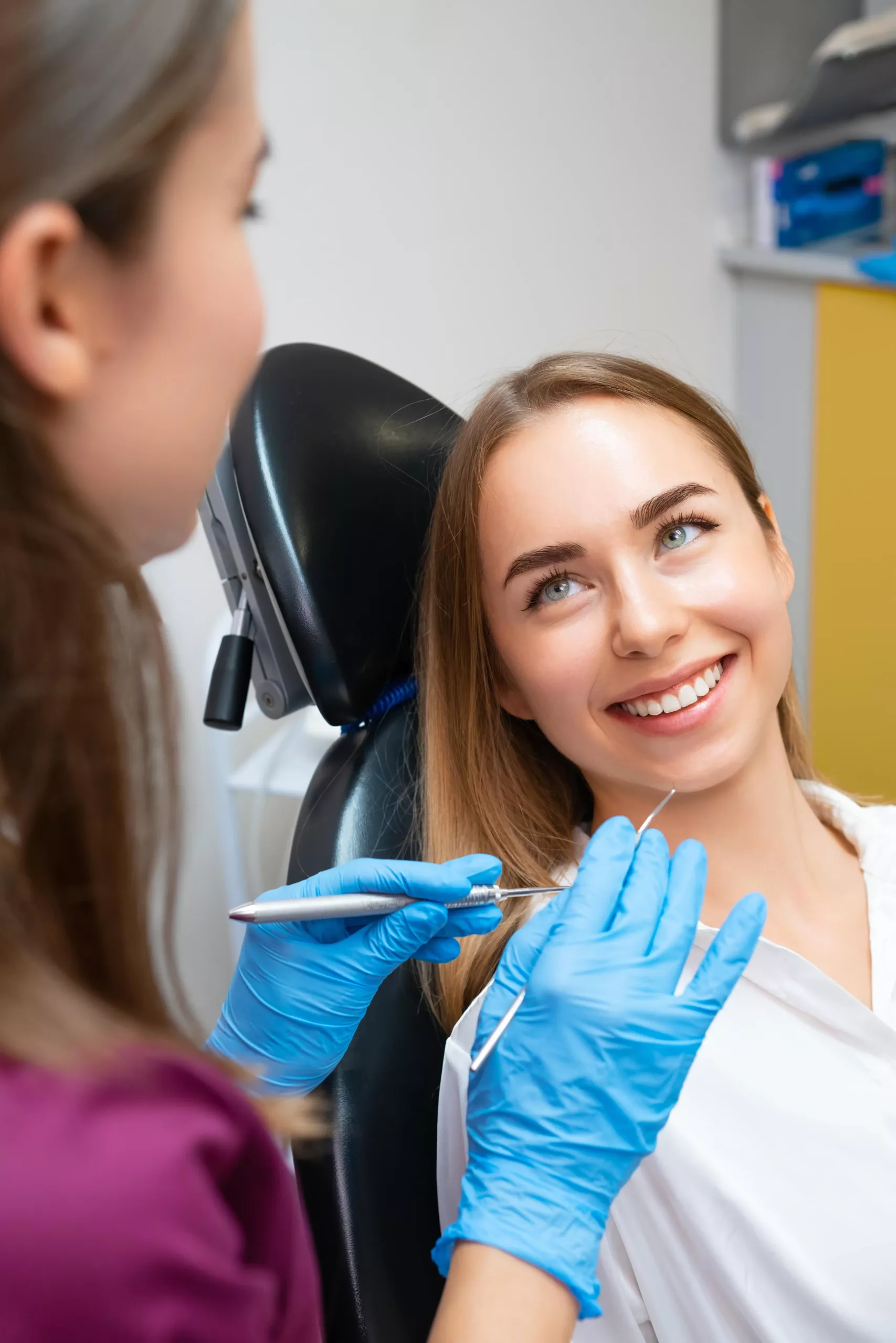 Belmont stock A woman is sitting in a dental chair, smiling up at a dentist wearing blue gloves who is holding dental tools.