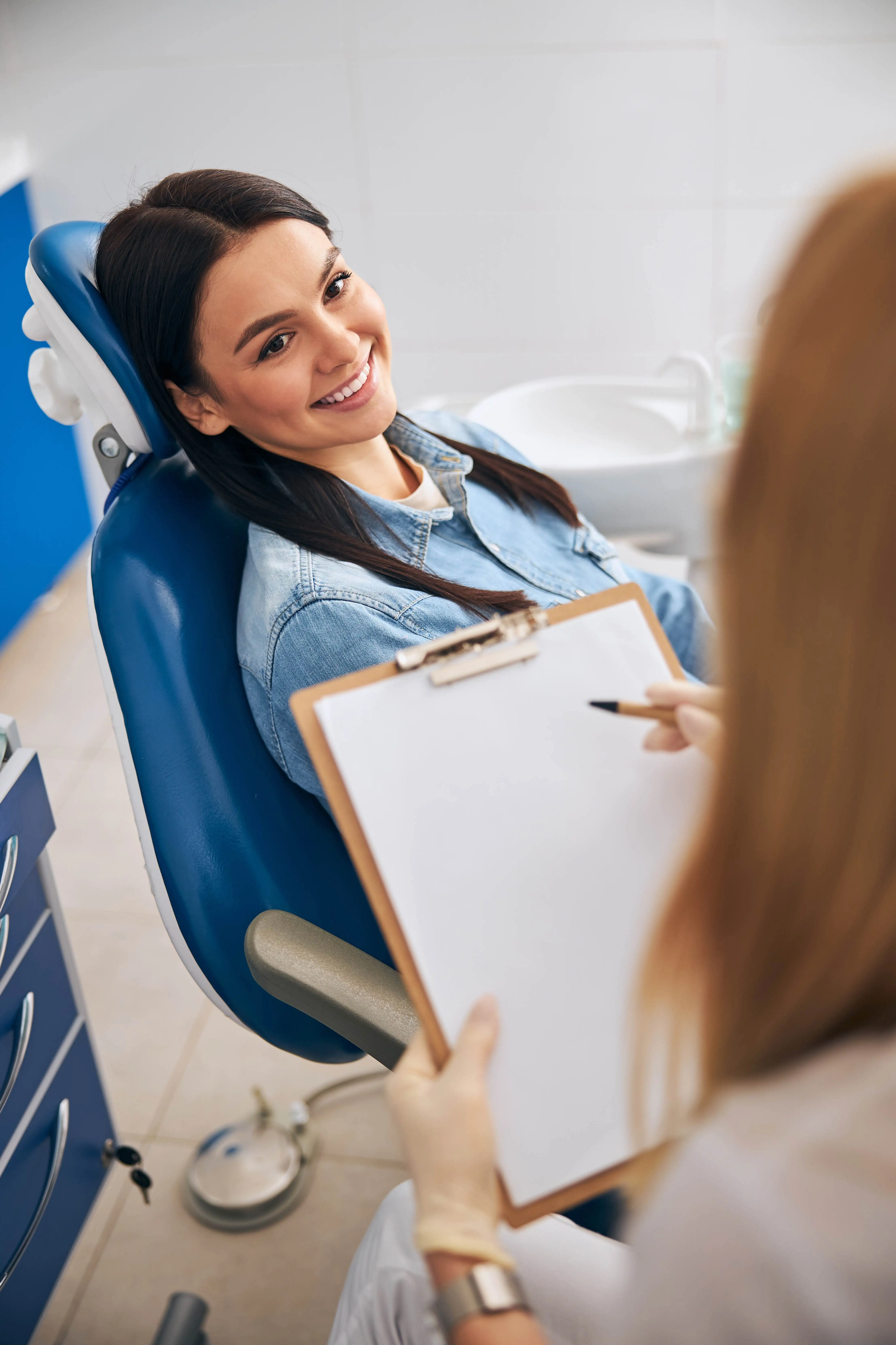 A woman in a dental chair smiles while a dental professional takes notes on a clipboard.