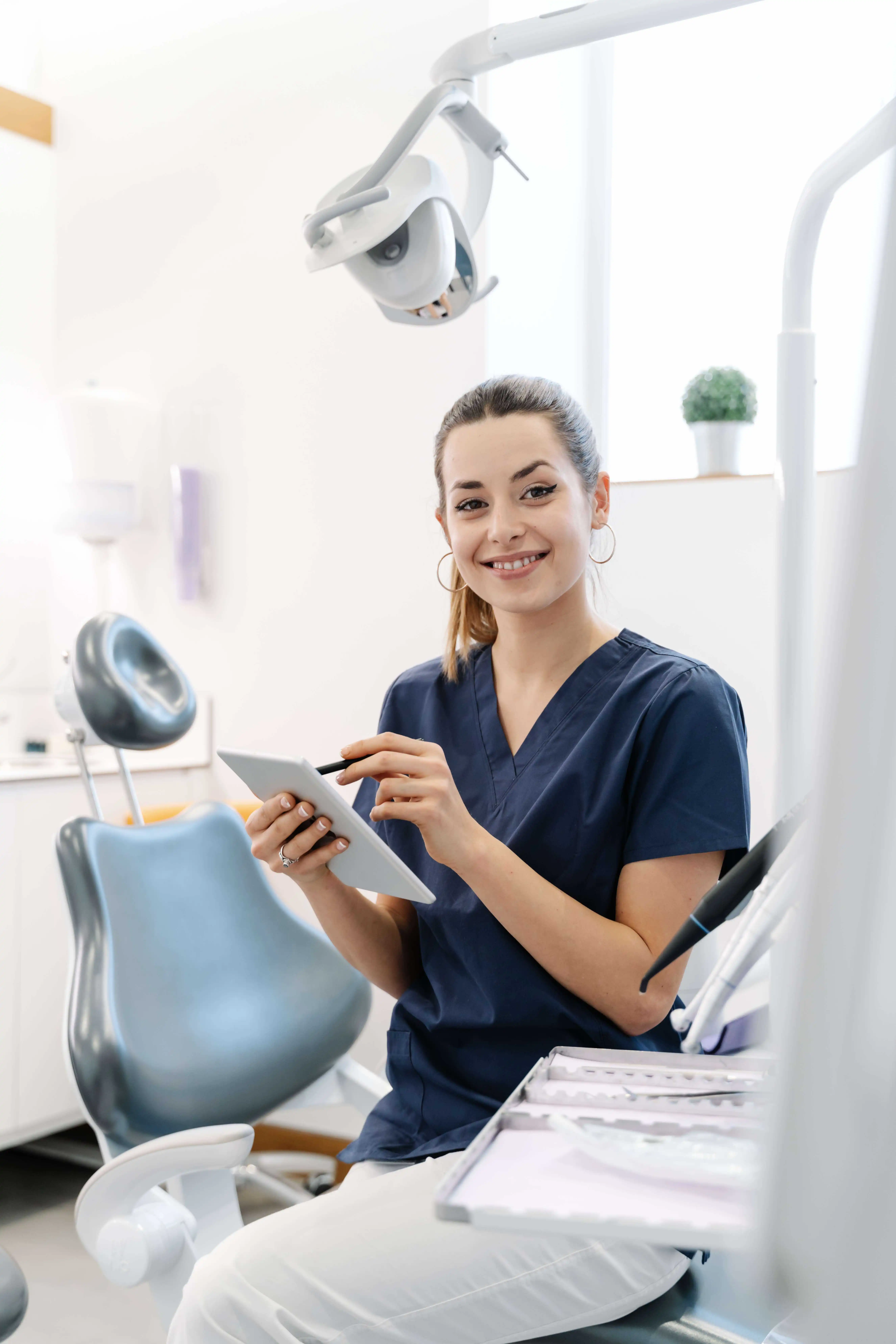 A woman in navy blue scrubs sits in a dental office, smiling and holding a tablet. Dental equipment and a chair are visible in the background.
