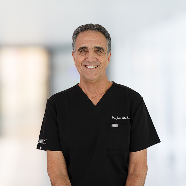 John Lapidus, DMD, standing and smiling, wearing black medical scrubs with a blurred, bright hallway in the background.
