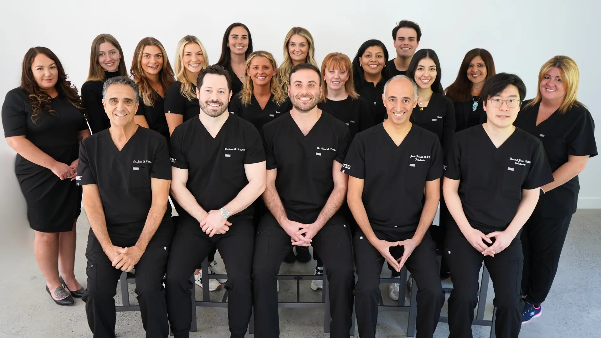 A group photo of 19 individuals, comprising both men and women, all wearing black uniforms. The group is smiling and posed in three rows against a plain white background.