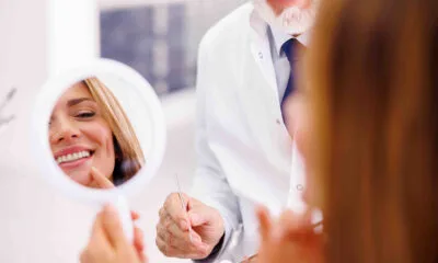 A woman looking at her reflection in a hand mirror, smiling, while a person in a white coat stands beside her.